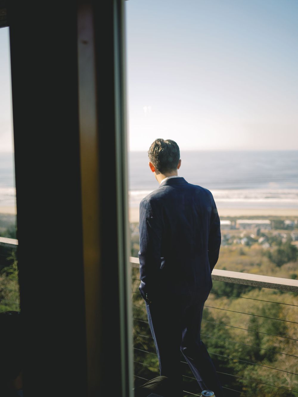 Groom in navy suit stands on balcony overlooking scenic coastal landscape on wedding day.