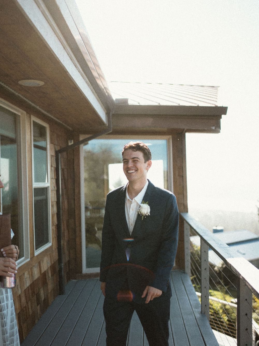 Smiling groom in navy suit with boutonniere standing on wooden deck at beach wedding.