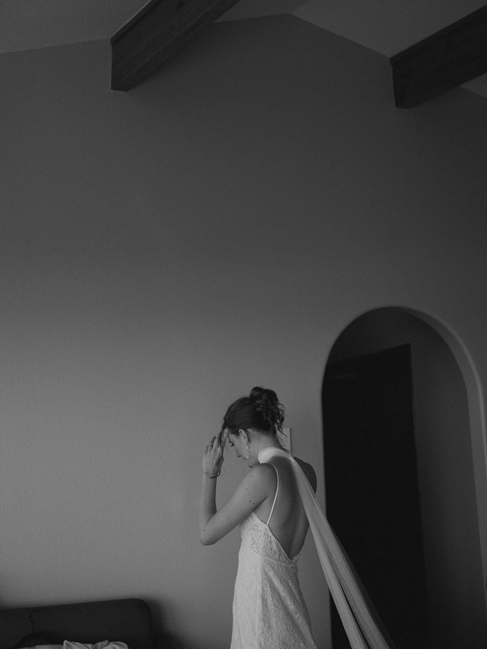 Black and white photo of bride in backless lace gown adjusting hair in room.