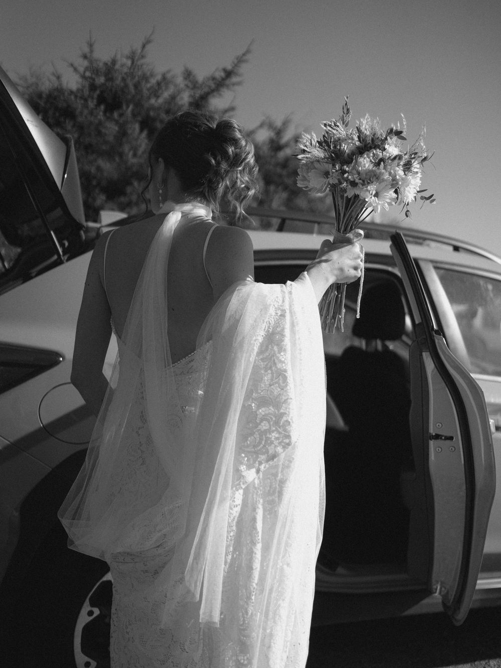 Black and white photo of bride in lace gown holding bouquet beside car door.