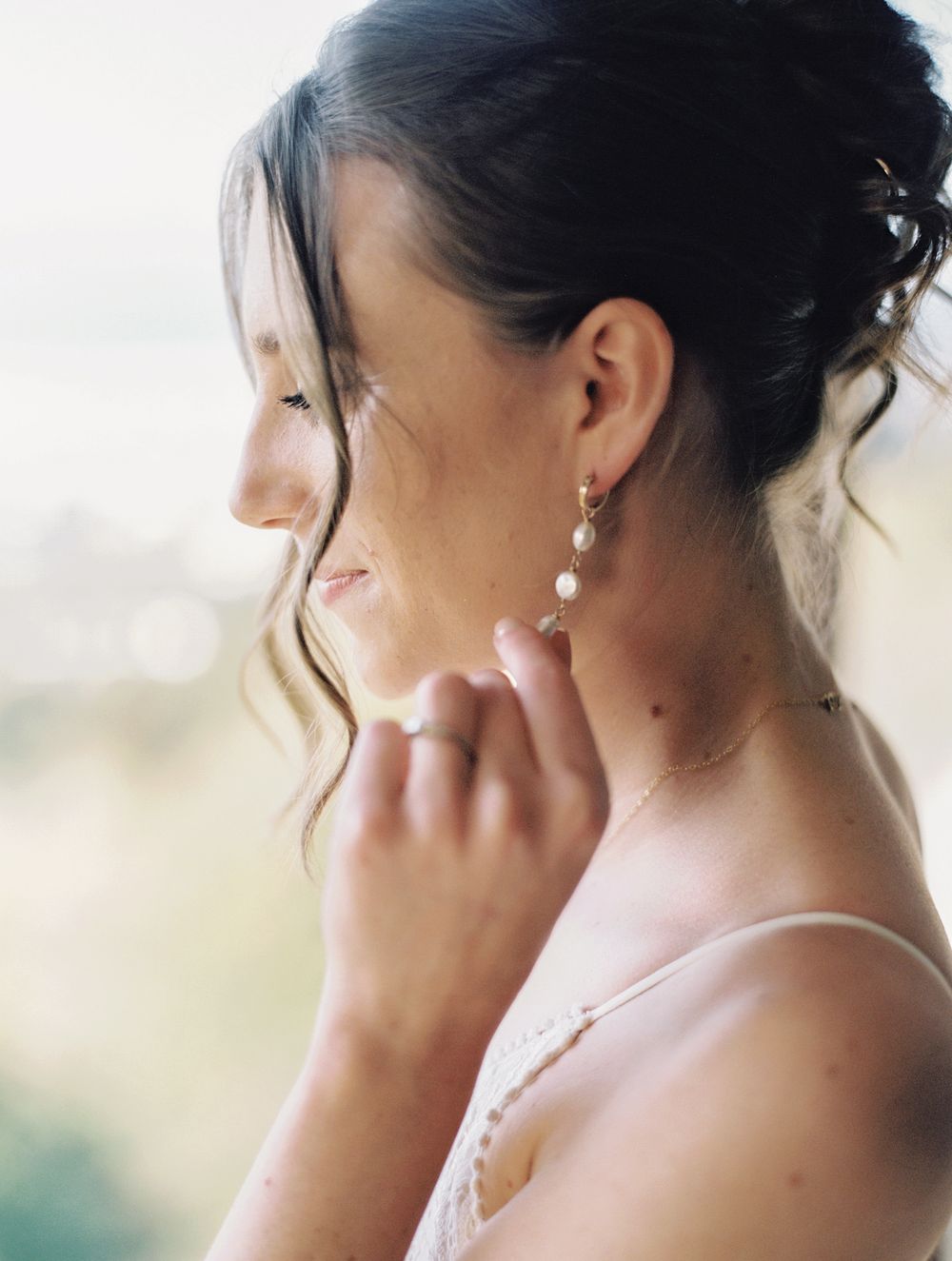 Close-up of a bride adjusting her pearl drop earrings, wearing a delicate lace wedding dress.