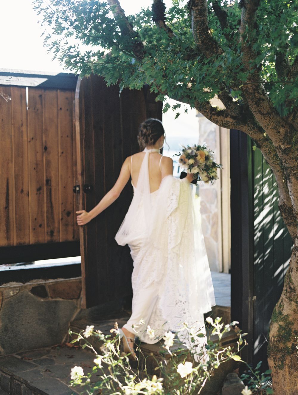 Bride in a white lace halter wedding gown holding a bouquet walking through a wooden garden gate.