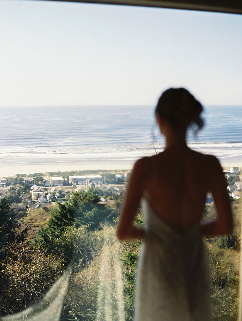 Bride in backless lace wedding dress gazing at ocean view through a large window.
