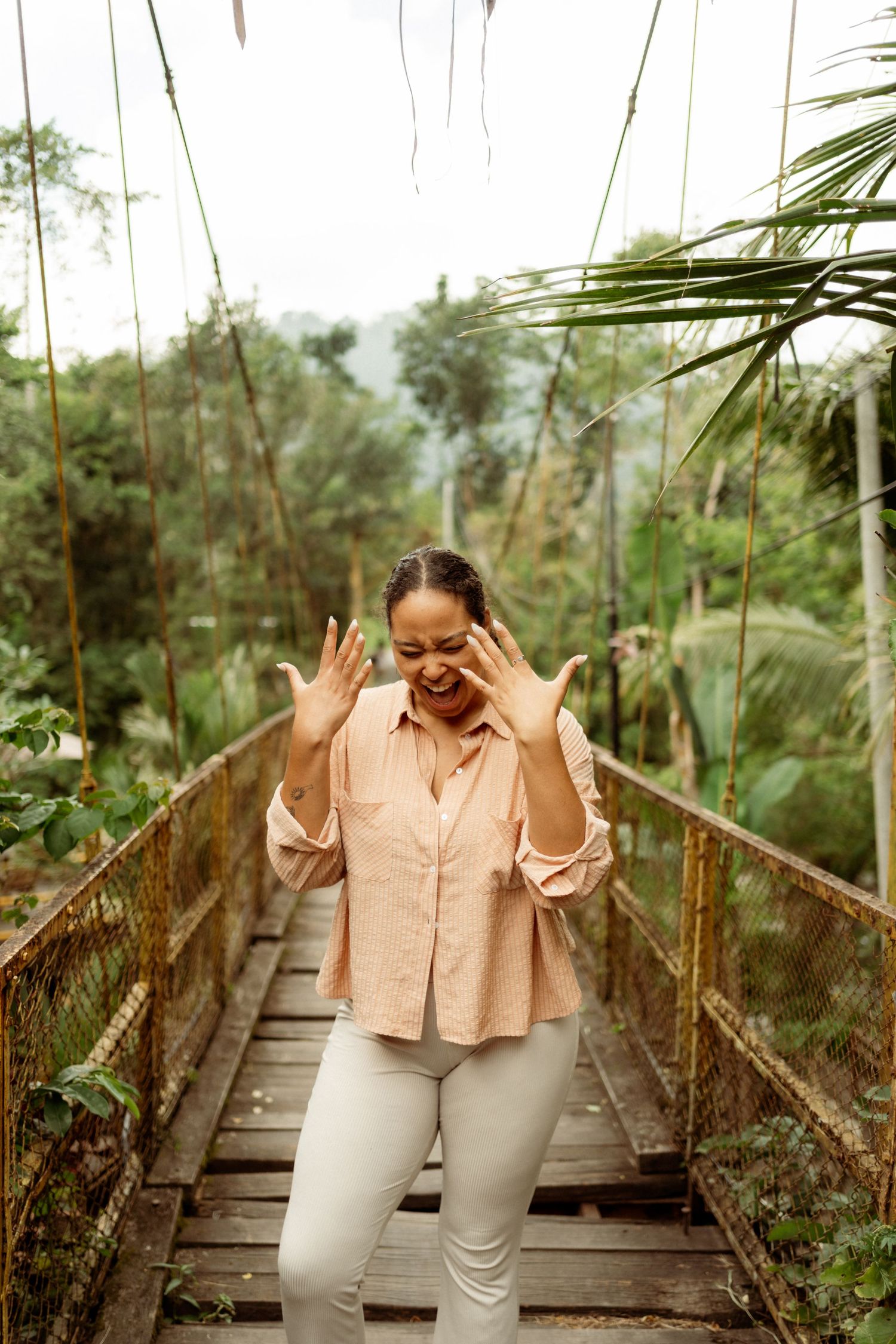 Woman in pink blouse laughing on a wooden suspension bridge surrounded by lush tropical jungle greenery.