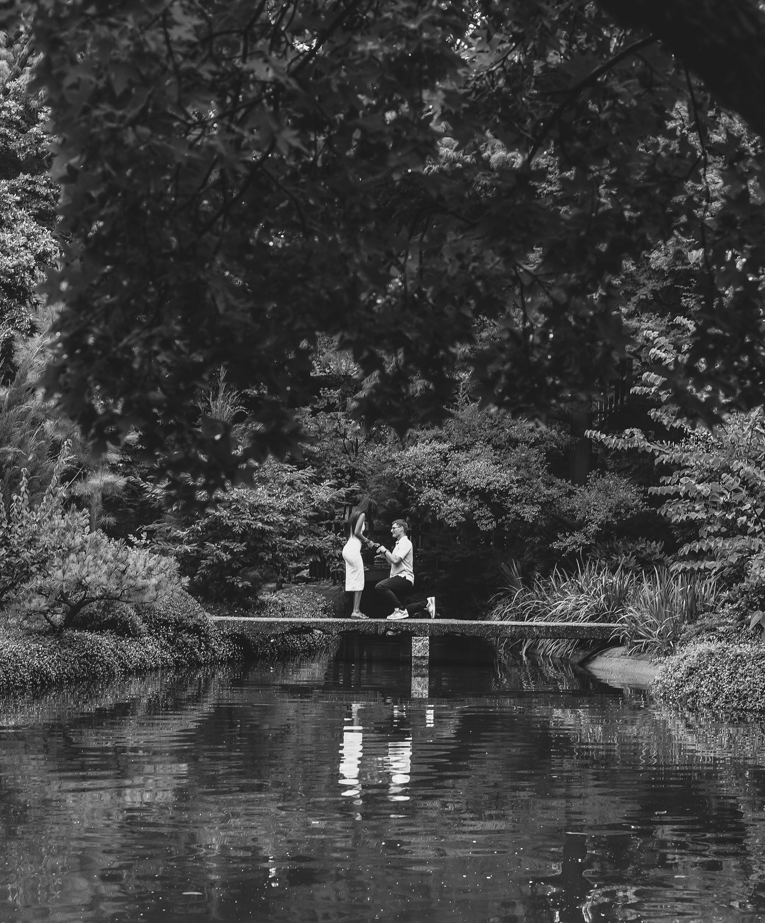 Black and white photo of two people standing on a dock surrounded by lush trees, reflected in a calm pond.