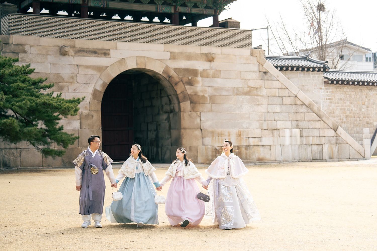 Four people in colorful traditional Korean hanbok hold hands walking near a stone gate at Gyeongbokgung Palace.