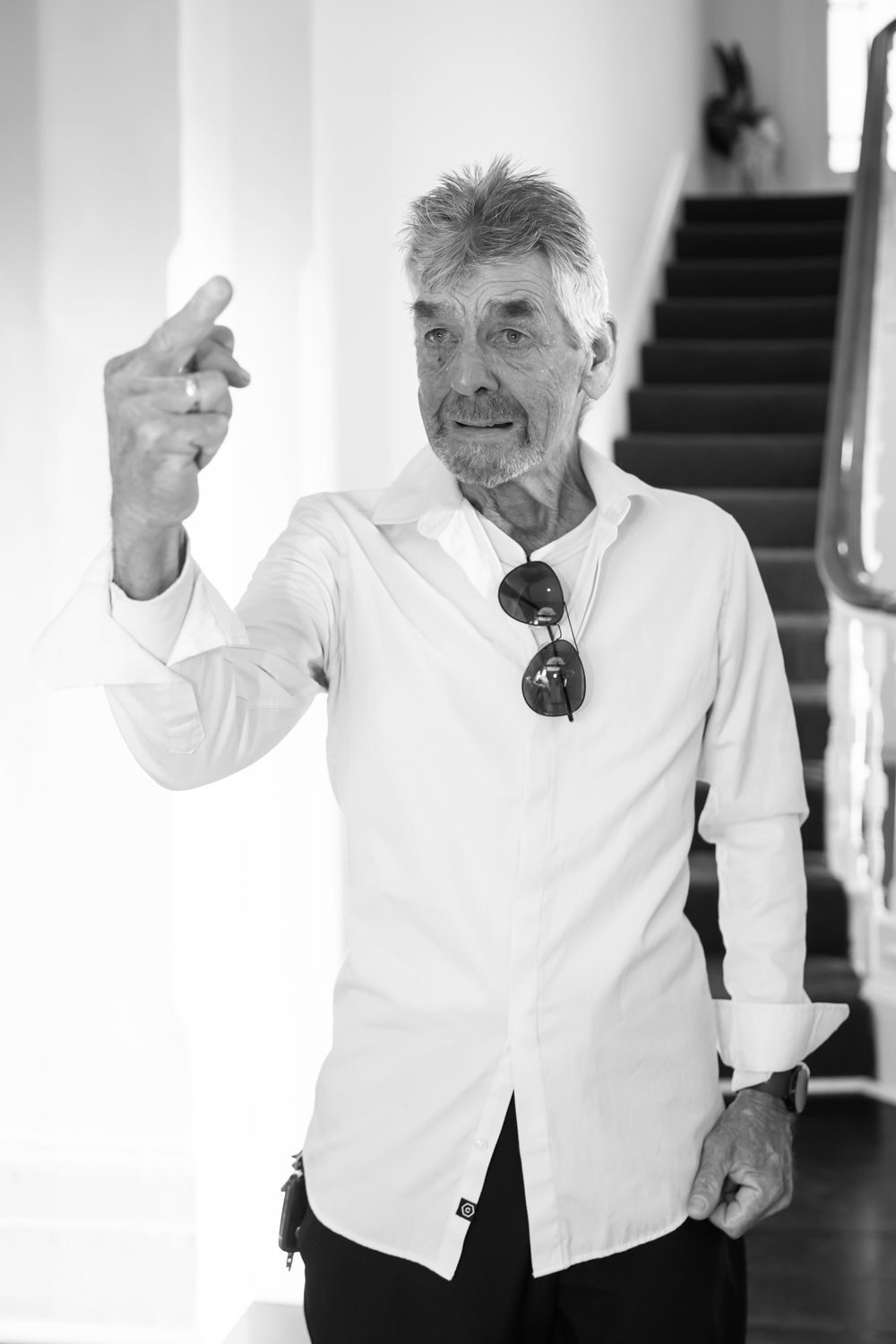 Black and white photo of an older man in white shirt with sunglasses, gesturing near a staircase.