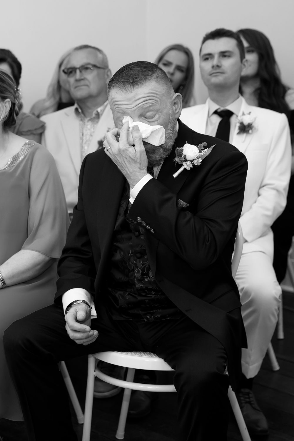 Emotional groom wiping tears with handkerchief during wedding ceremony, wearing black suit with boutonniere, black and white photo.
