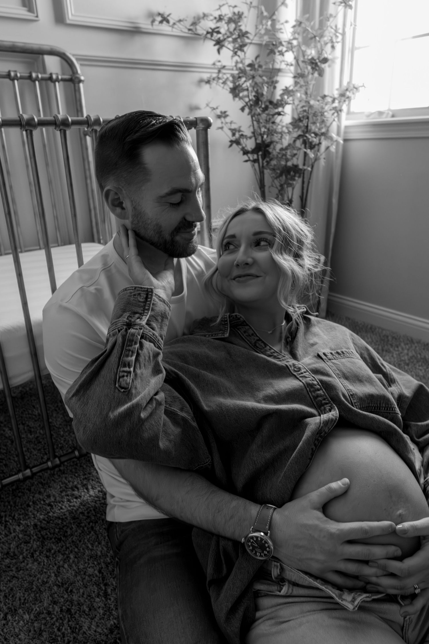 Black and white maternity photo of couple sitting together on floor, man embracing pregnant woman.