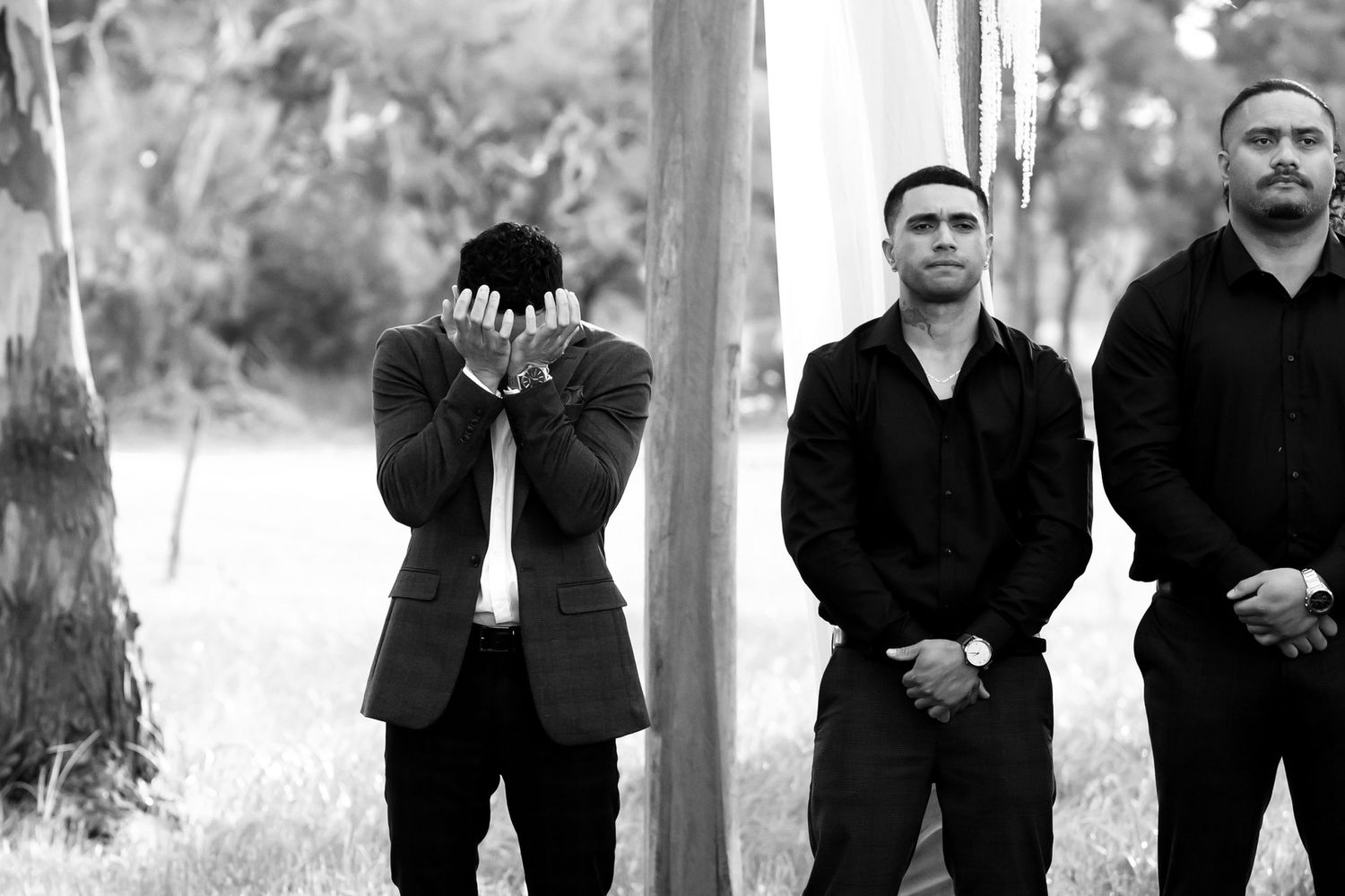 Three men in black formal attire stand outdoors, one covering his face with hands in an emotional moment.