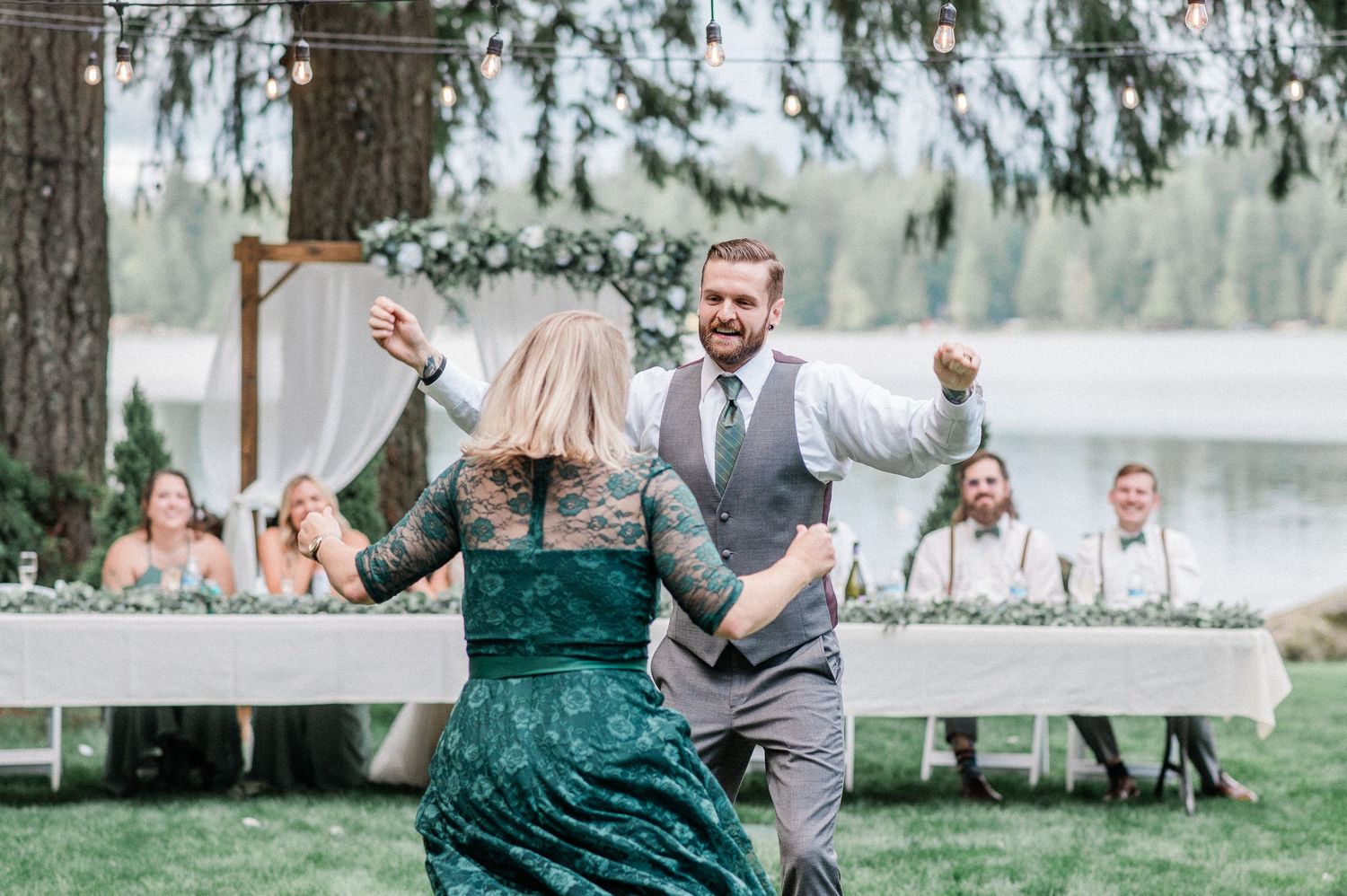 Groom dancing with mother in green lace dress at outdoor wedding reception with string lights.