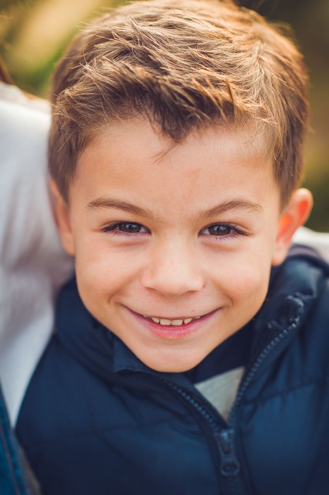 Smiling young boy with brown hair wearing a navy blue jacket, photographed outdoors in warm natural light.