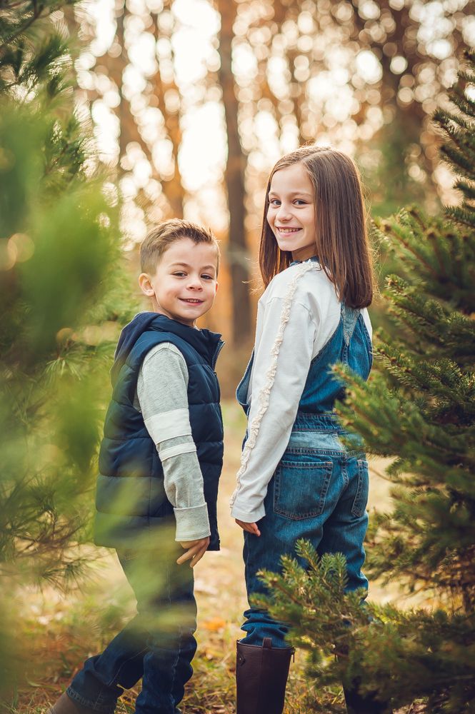 Two smiling children pose among pine trees in golden autumn light, girl in denim overalls and boy in navy vest.