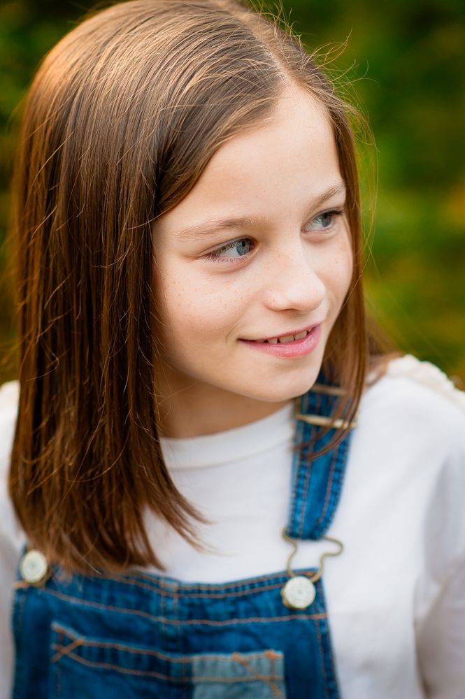 A young girl with brown hair wearing denim overalls smiles softly outdoors with green foliage in the background.