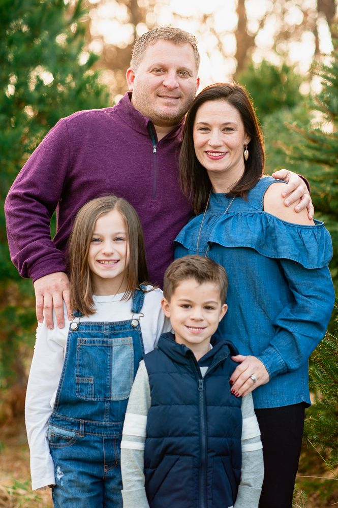 Happy family of four posing outdoors among pine trees, parents with two young children in casual fall clothing.