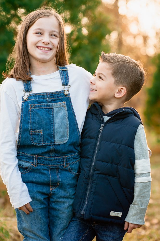 Happy girl in denim overalls and boy in navy vest share a joyful moment together outdoors in warm sunlight.
