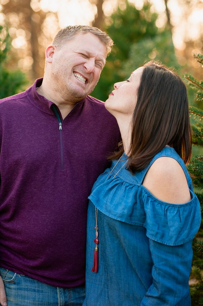 A laughing couple poses outdoors near evergreen trees, woman in blue off-shoulder top leans toward smiling man in purple pullover.