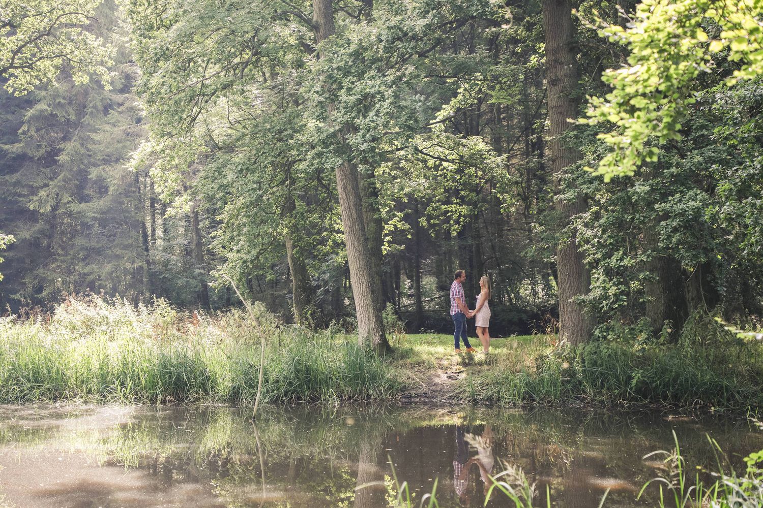 Couple holding hands by a tranquil forest pond surrounded by tall trees and lush green foliage on a sunny day.