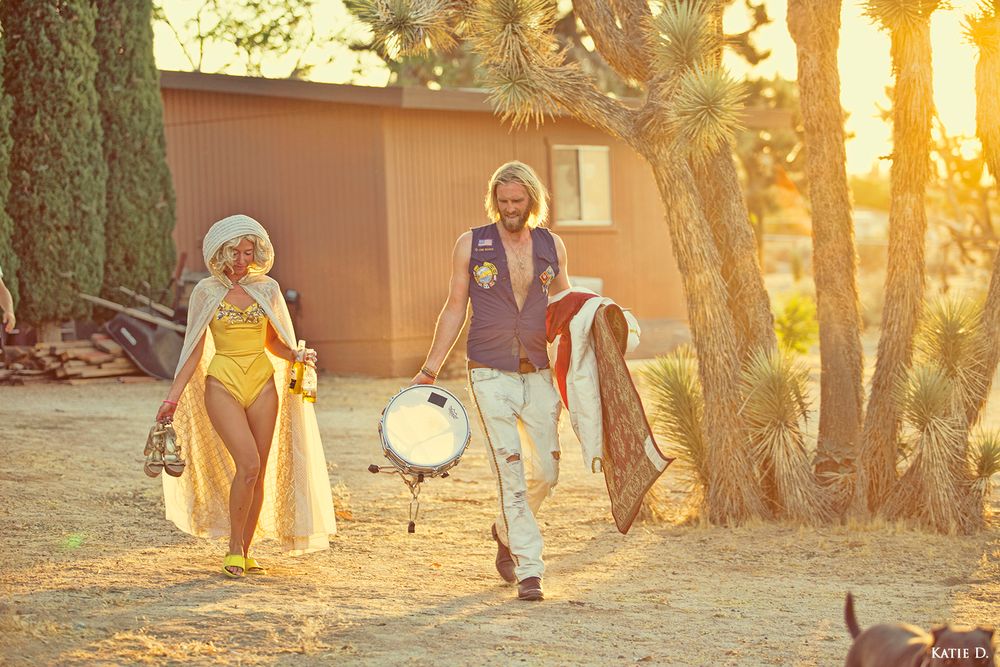 Two musicians carrying drum and guitar walking through desert landscape with Joshua trees at sunset.