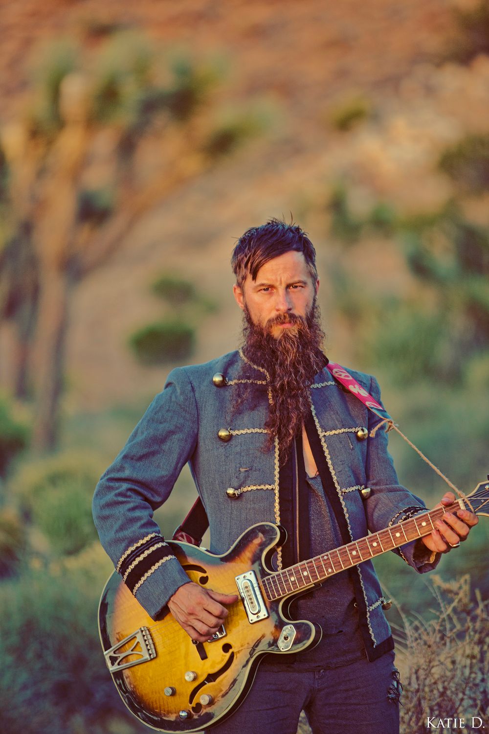 Bearded male guitarist holding a sunburst hollow-body electric guitar in Joshua Tree National Park desert.