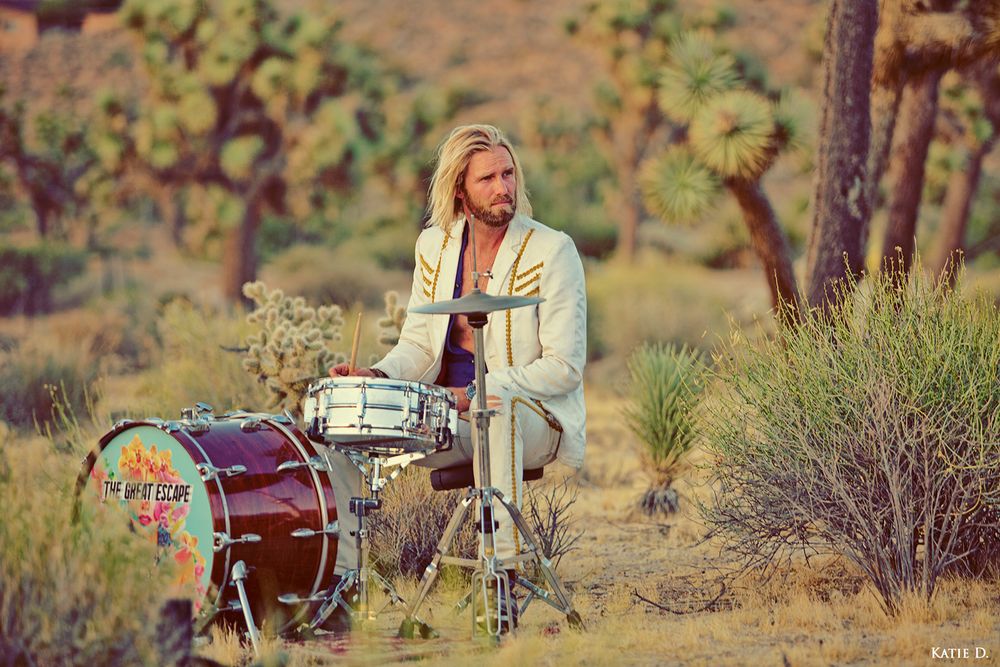 Blonde male drummer playing a drum kit outdoors in a Joshua Tree desert setting at sunset.