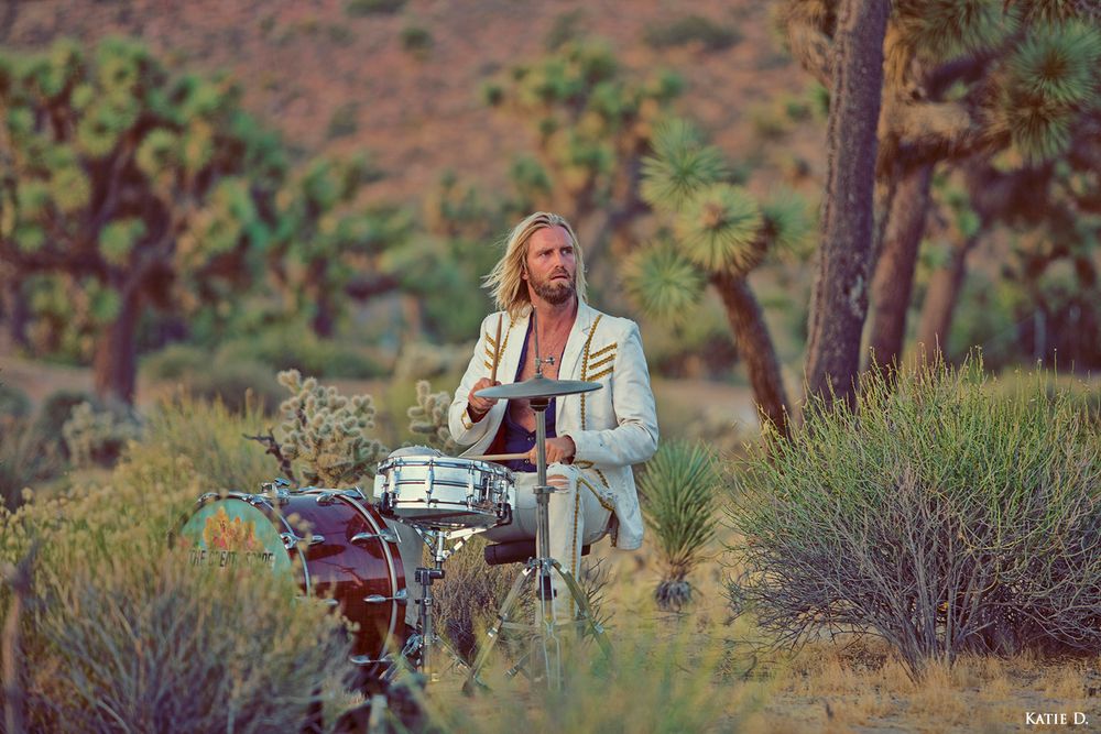 Long-haired blonde drummer in a white jacket playing drums in a scenic Joshua Tree desert landscape.