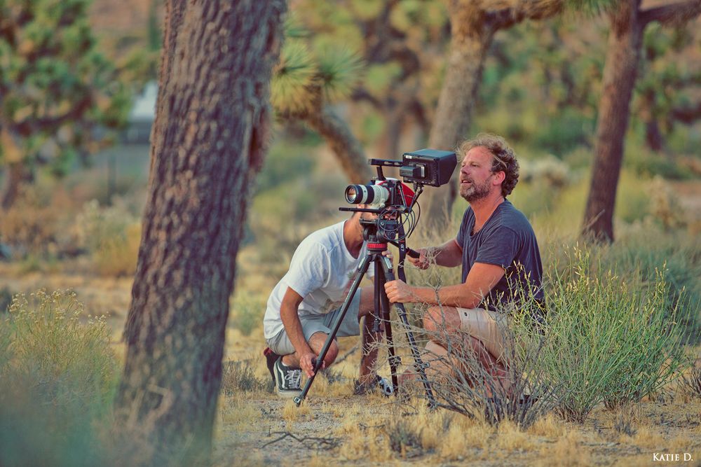 Two filmmakers set up a camera on tripod among Joshua trees in a desert landscape.