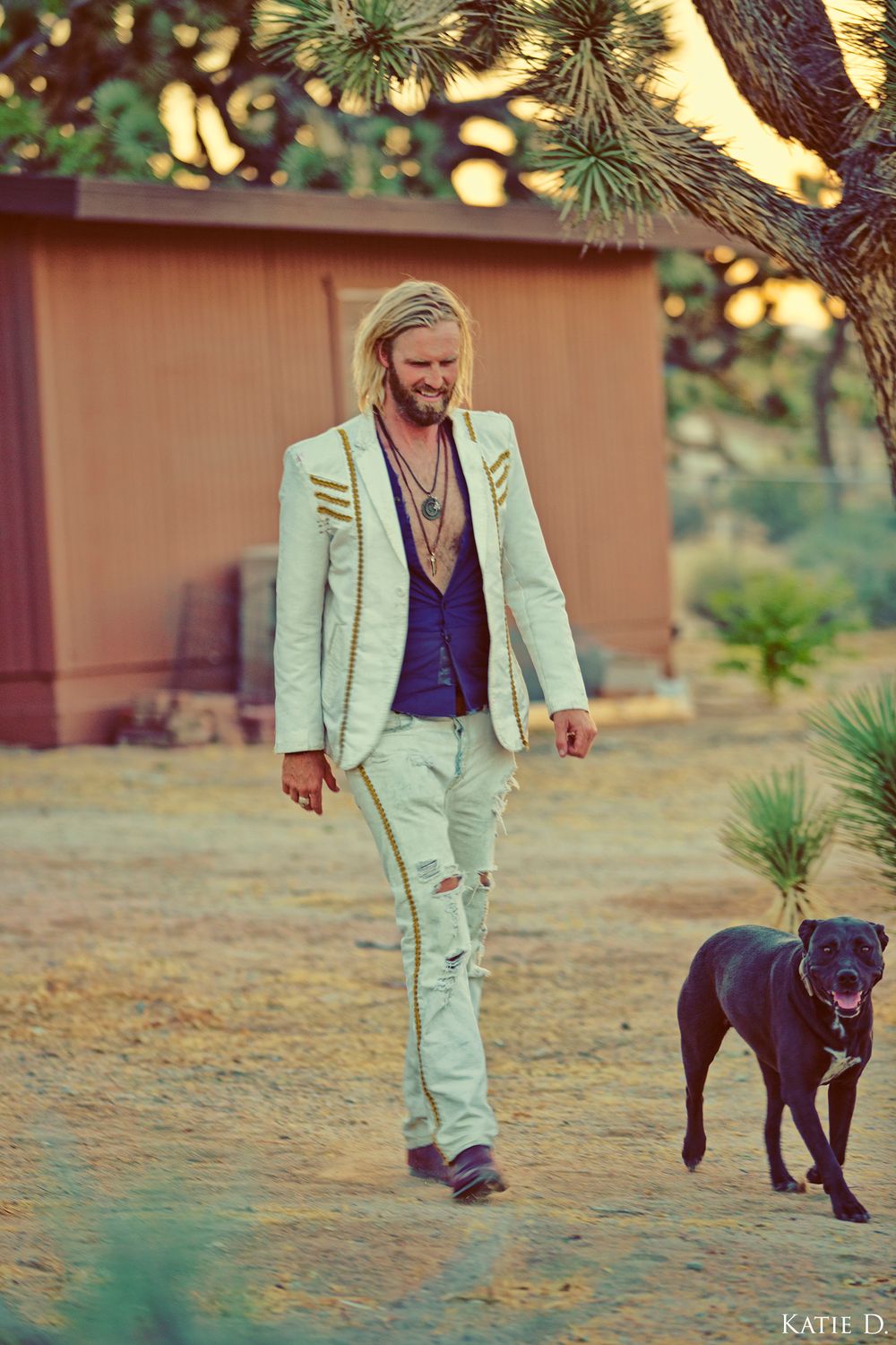 Blonde man in stylish white blazer walks with a black dog in a desert setting.