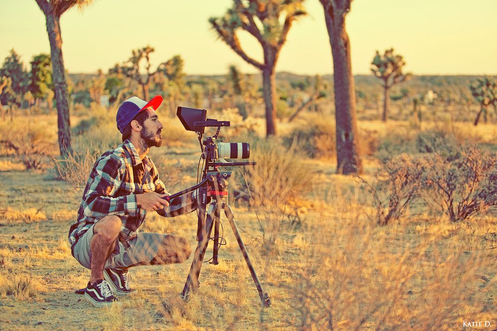 Bearded male photographer crouching behind camera on tripod in Joshua Tree desert.