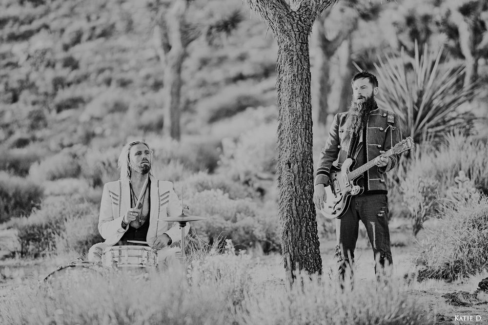 Black and white photo of two musicians performing with drums and guitar in a desert Joshua tree landscape.