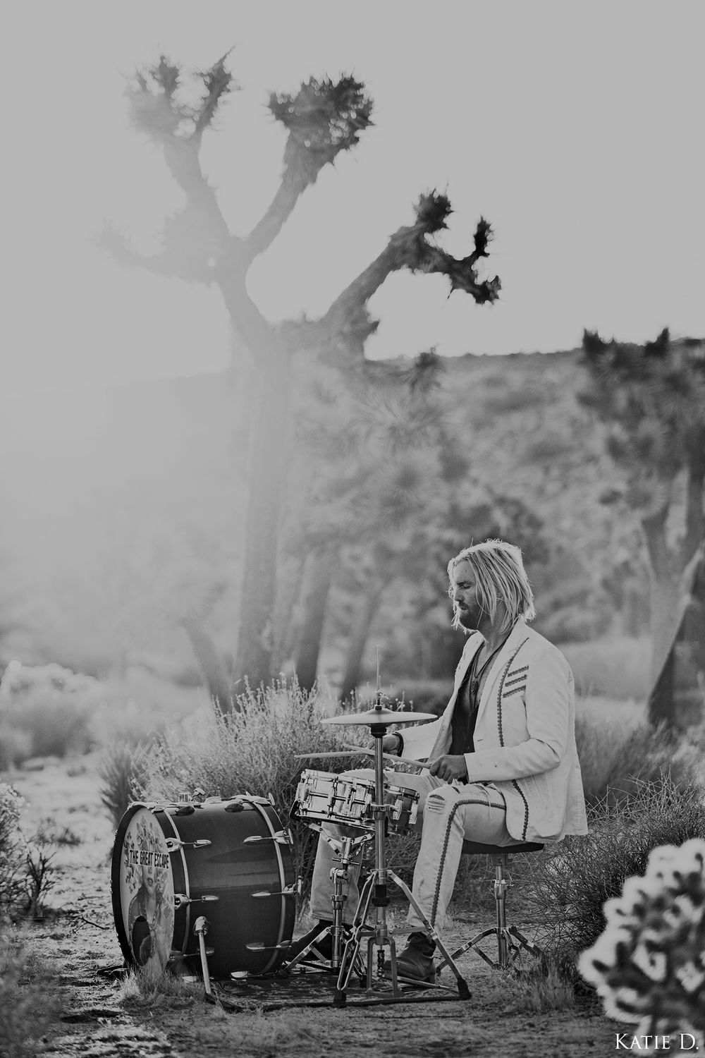 Black and white photo of a bearded drummer in a white suit playing drums beside a Joshua tree.