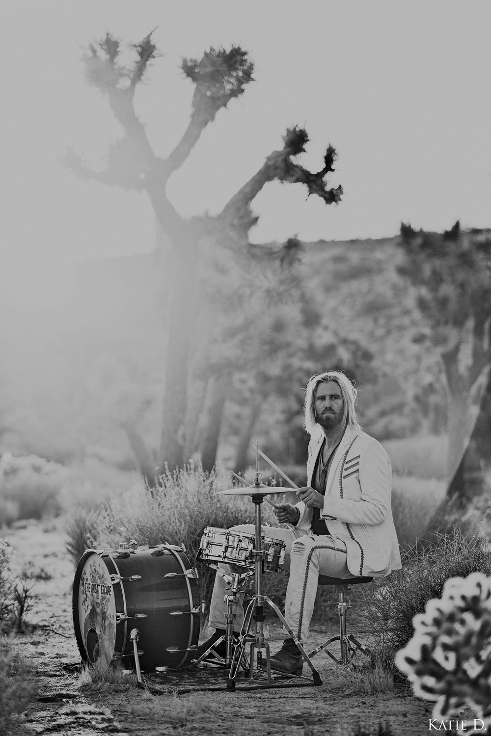 Black and white portrait of a bearded drummer playing a drum kit in a misty desert Joshua tree setting.