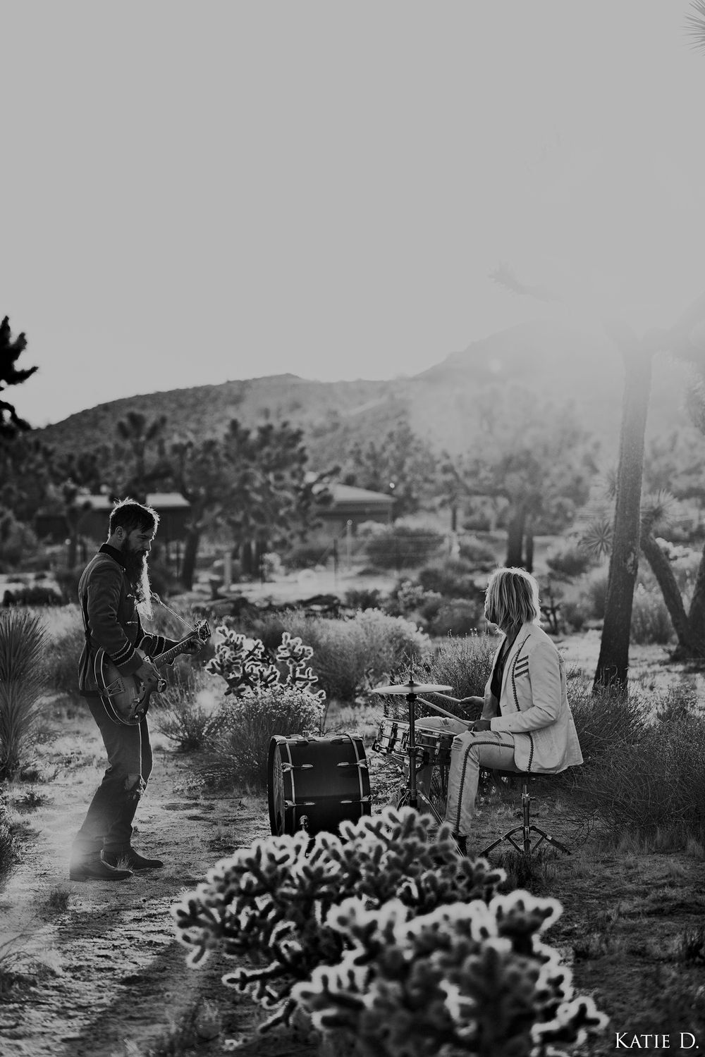Black and white photo of two musicians playing guitar and drums in a desert landscape with Joshua trees.