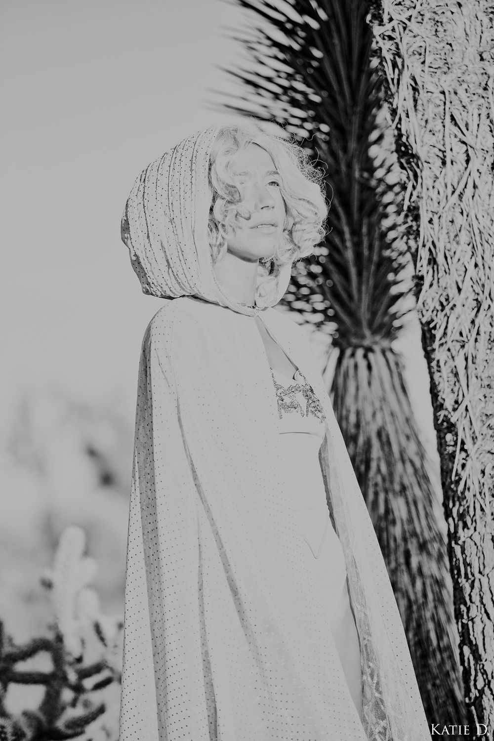 Black and white portrait of a woman in a hooded cape standing next to a tree.