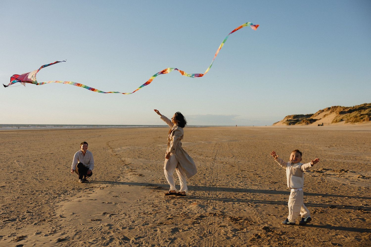 Family flying colorful kites on a sunny beach with sand dunes, enjoying a fun outdoor activity together.