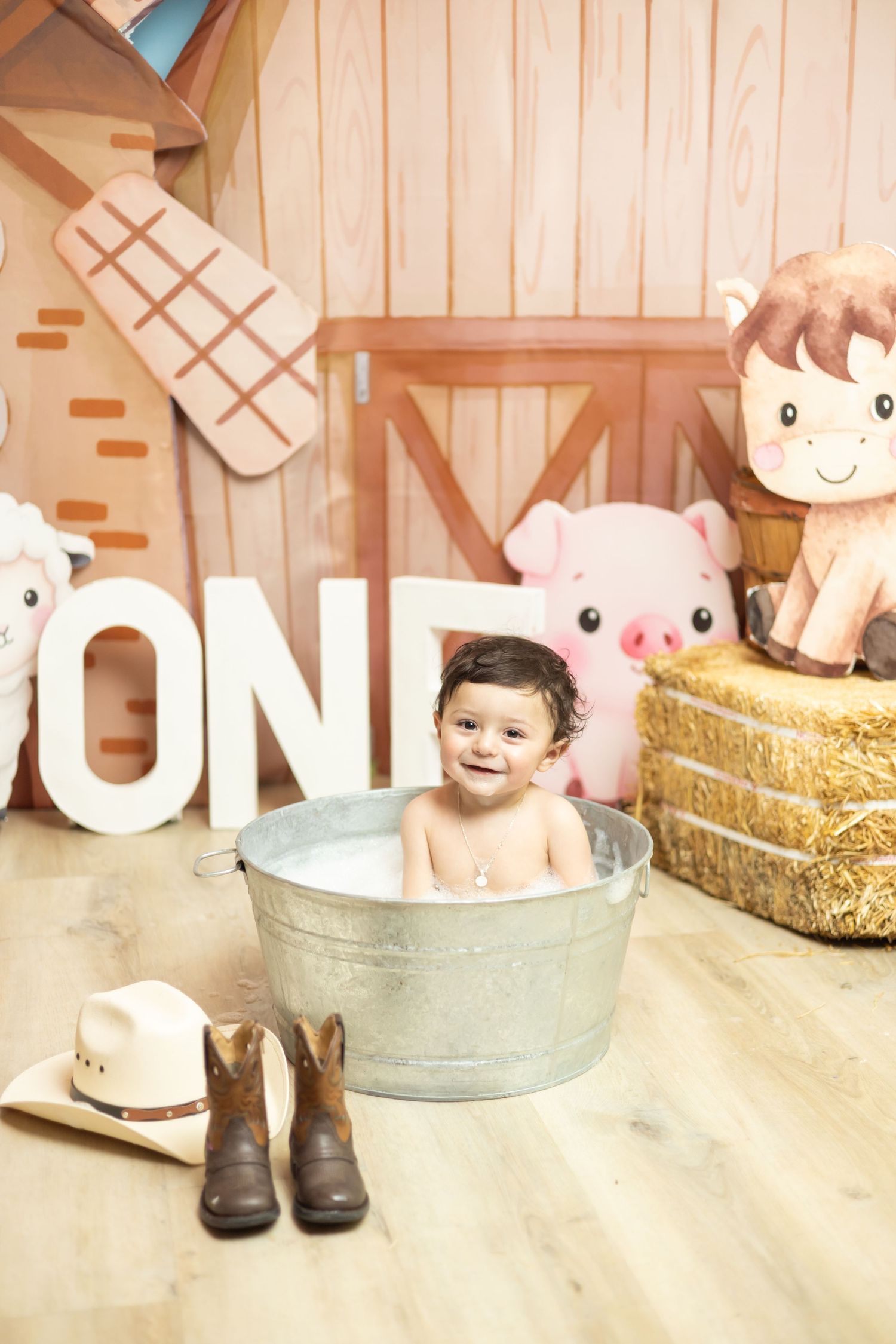 Baby sitting in galvanized tub at farm-themed first birthday party with hay bales, animal toys, and cowboy boots.
