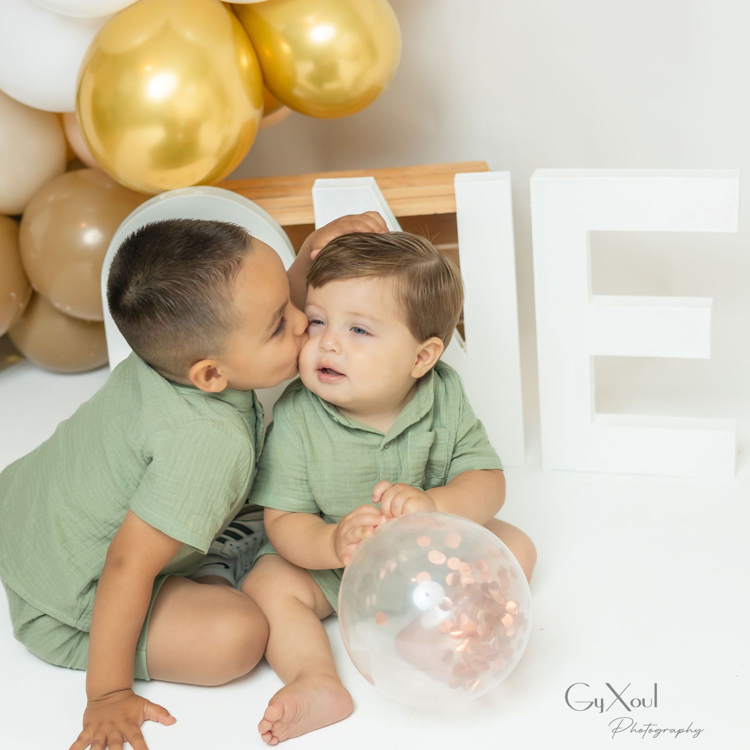 Two toddlers in matching green outfits share a sweet kiss near gold balloons and a white letter E decoration.