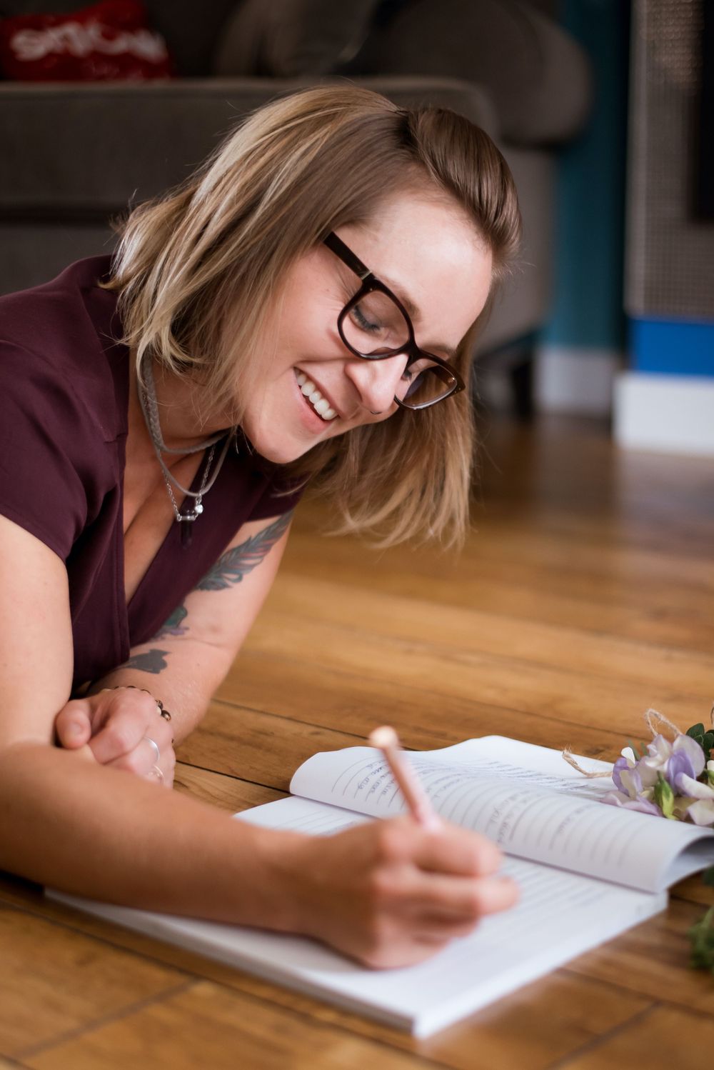 Smiling woman with glasses writing in a notebook while lying on a hardwood floor.