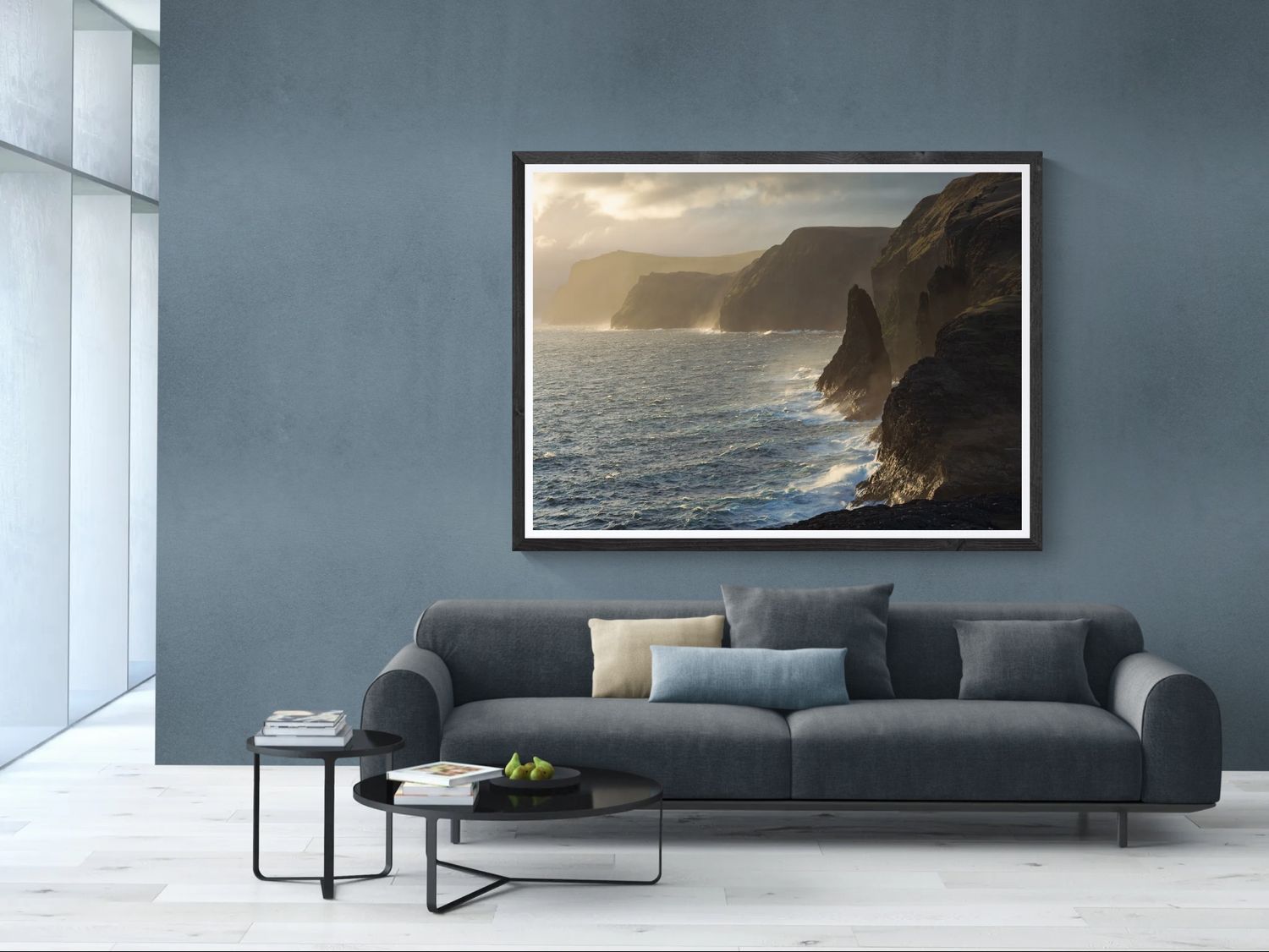 Framed coastal cliff landscape photograph hanging above a modern gray sofa in a blue living room.