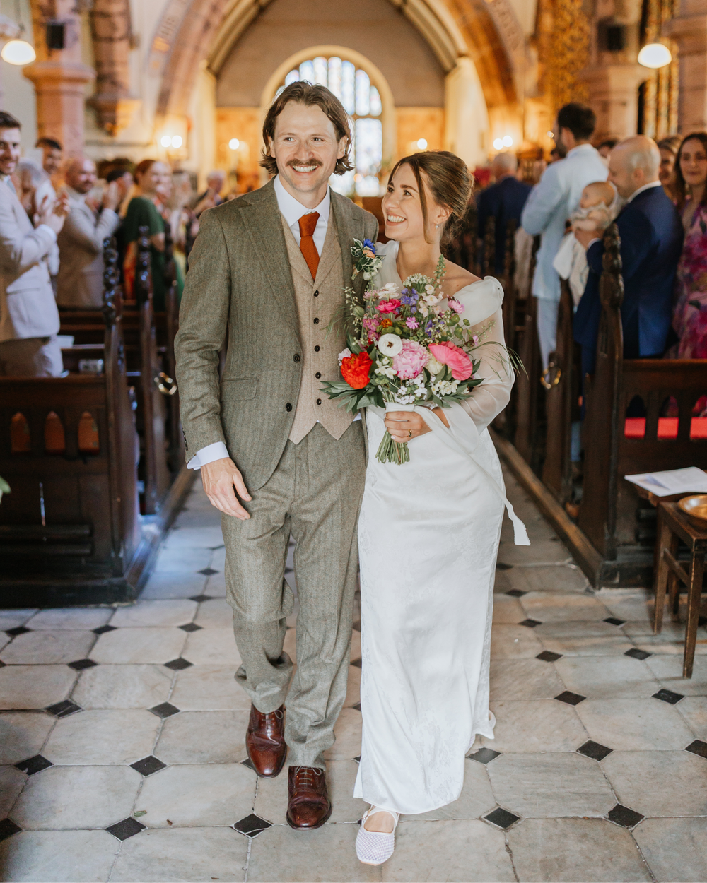 A joyful bride and groom walk down the aisle of a church, she holds a colorful bouquet, guests applaud in the background.