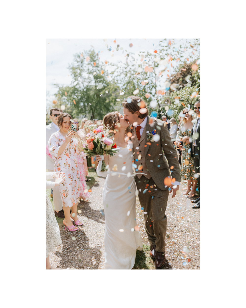 Newlyweds share a kiss while colorful confetti fills the air, surrounded by cheering guests at an outdoor wedding.
