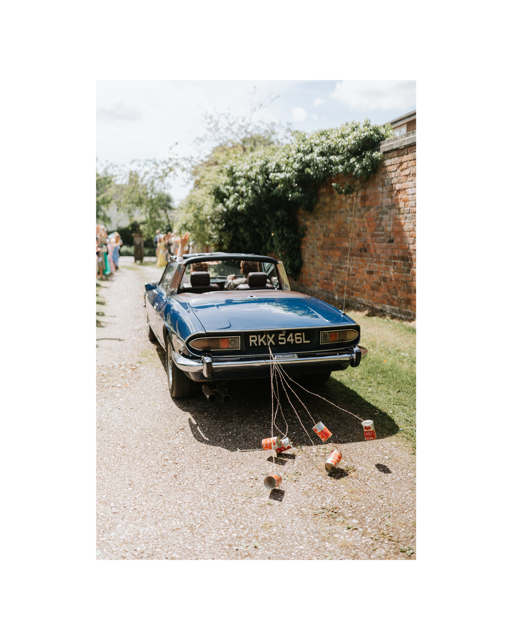 Blue vintage wedding car with tin cans tied to the back bumper, parked on a gravel driveway beside a brick wall.
