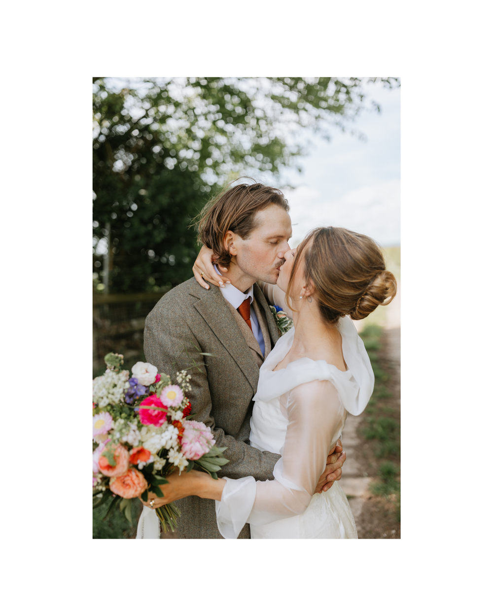 A bride and groom share a romantic kiss outdoors, she holds a vibrant bouquet of pink and orange flowers.