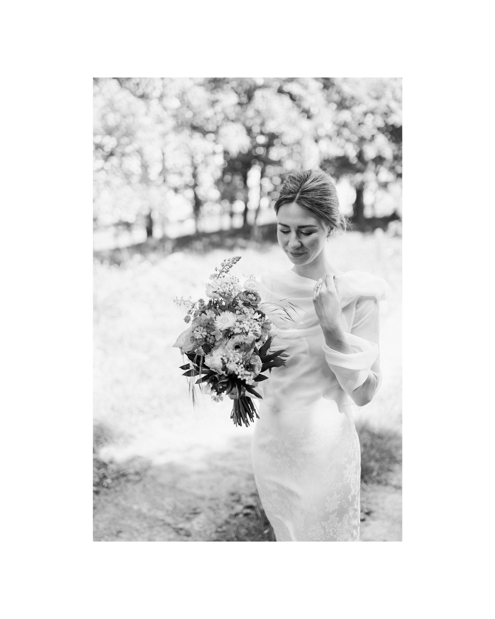 Black and white photo of a bride in a elegant gown holding a floral bouquet outdoors among trees.