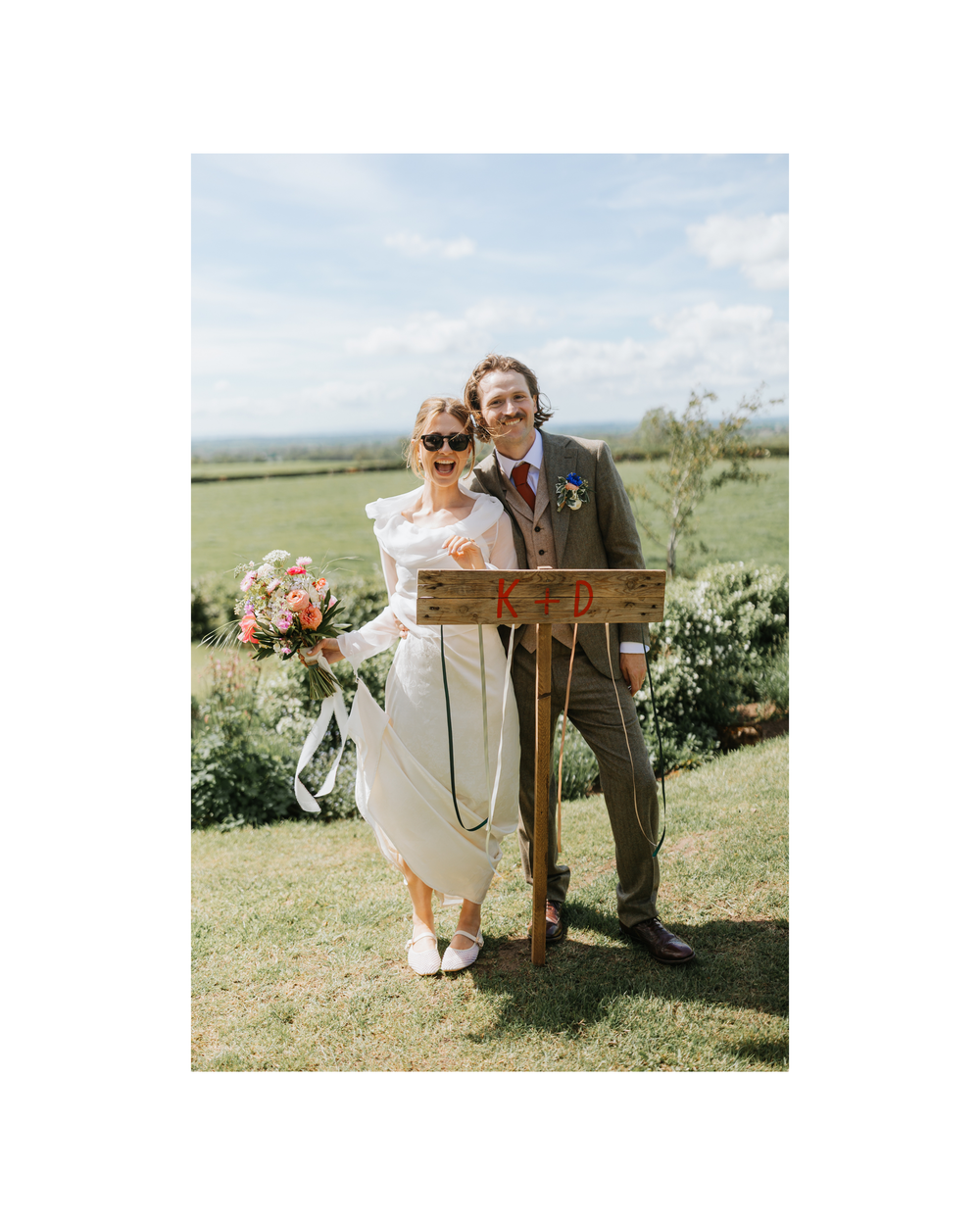 Happy couple posing outdoors at a rustic countryside wedding, bride holding colorful bouquet wearing sunglasses, groom in tweed suit.