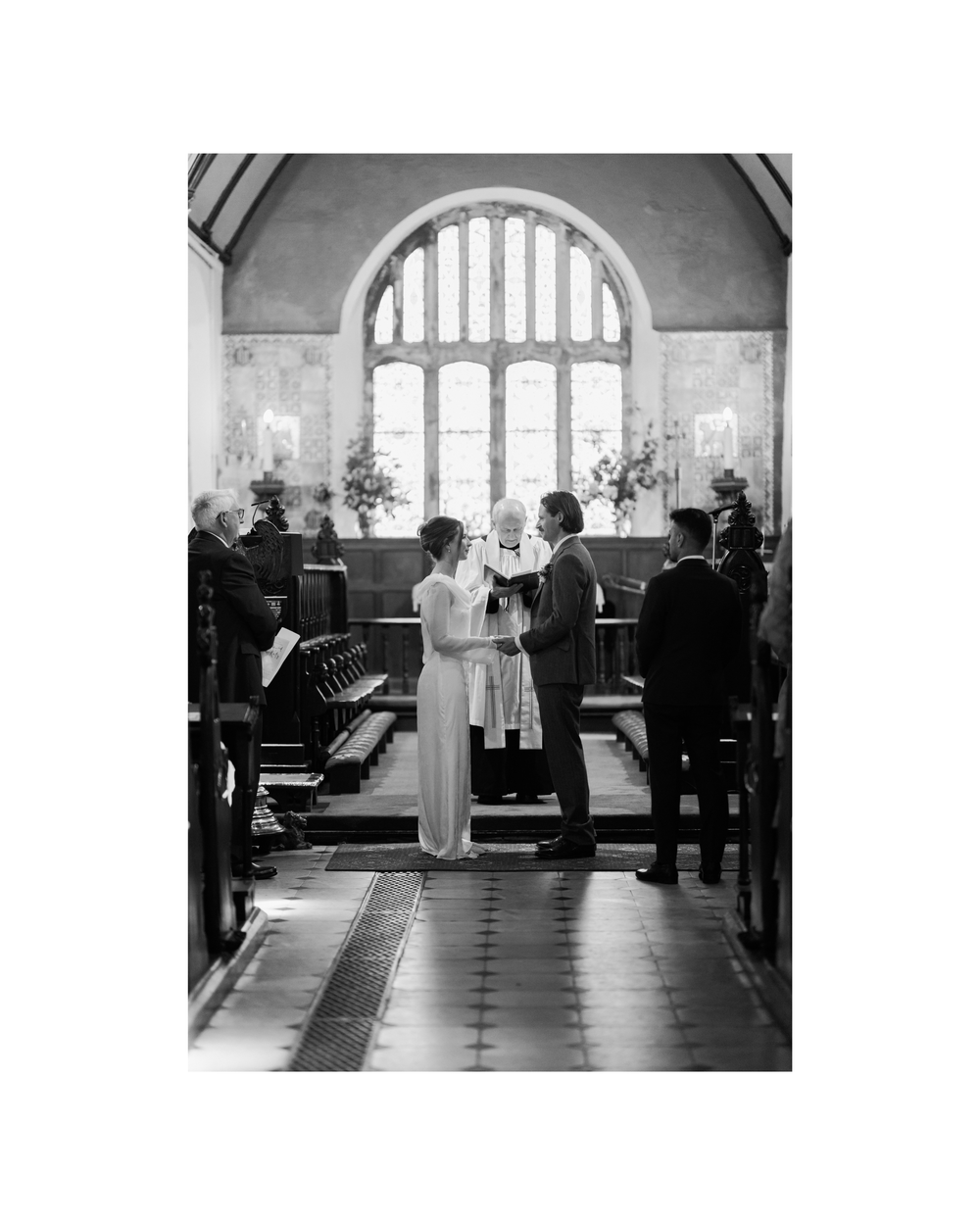 Black and white photo of a couple exchanging vows at a church altar with an officiant and arched window backdrop.