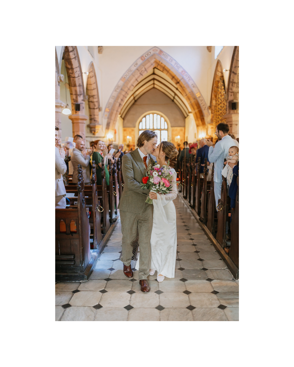 Bride and groom share a kiss in a beautiful stone church aisle, surrounded by guests, bride holding a colorful bouquet.