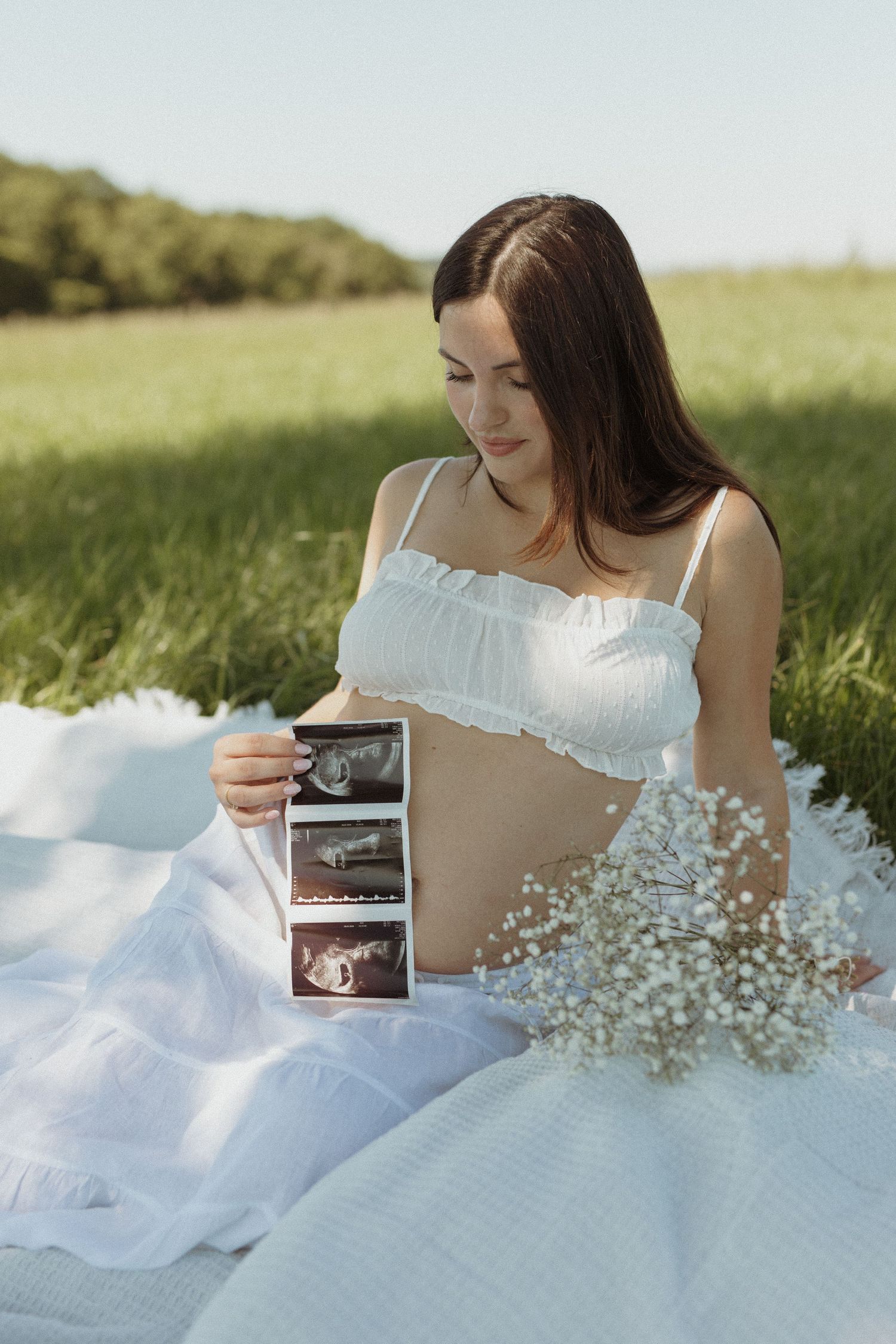 Pregnant woman in white outfit sits on blanket in green field holding ultrasound photos.
