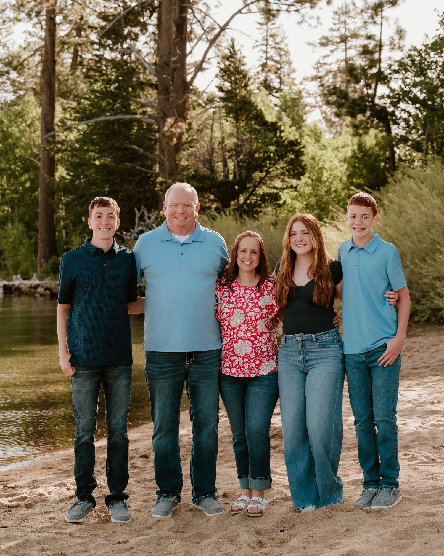 Family of five smiling together on a sandy beach surrounded by trees in a warm outdoor portrait.