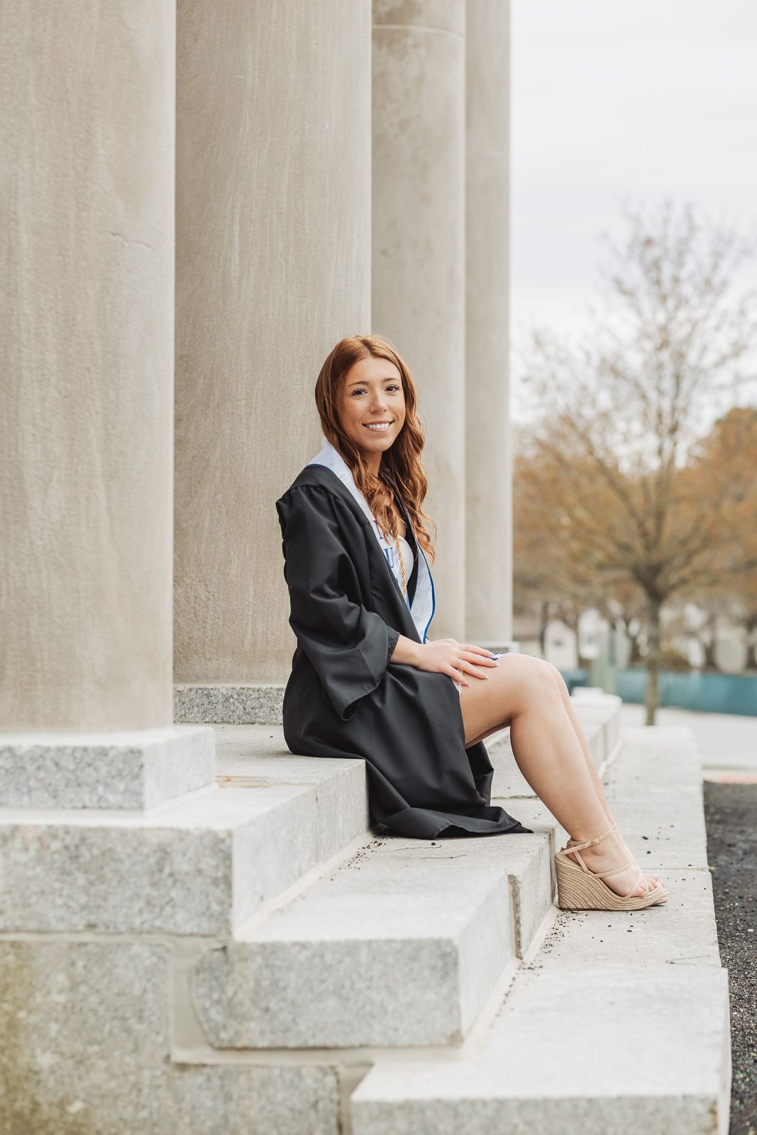 Smiling female graduate in black cap and gown sitting on stone steps between large columns at university.
